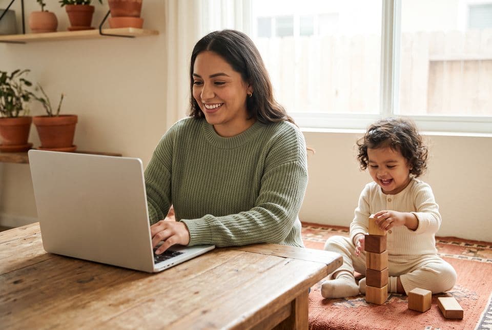 A young mother working on a laptop at a kitchen table while her toddler plays with wooden blocks beside her